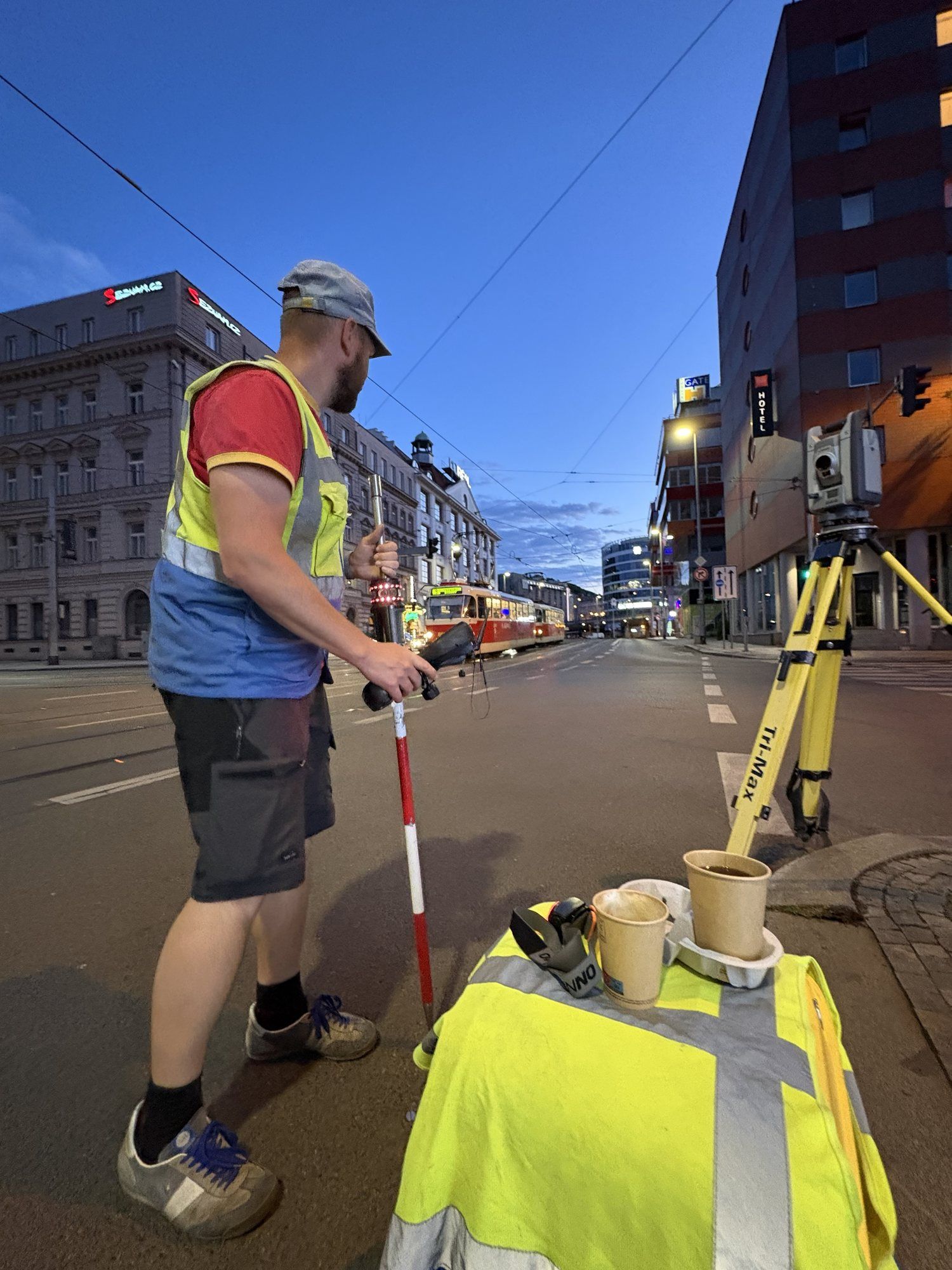 Geodet v reflexní vestě s výtyčkou a ovladačem uprostřed prázdné ulice s tramvají, vpravo totální stanice na žlutém stativu