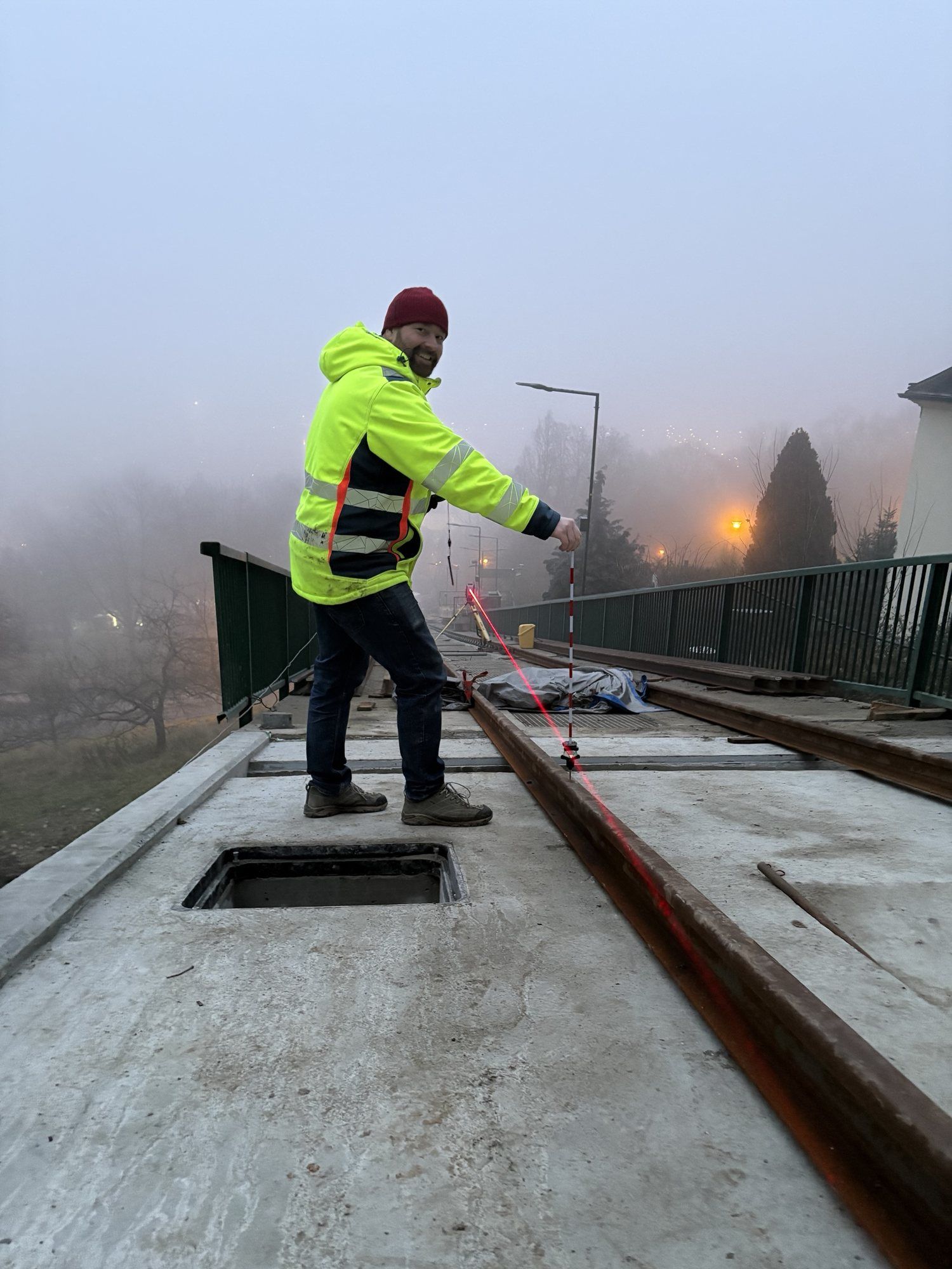 Usměvavý geodet v reflexní bundě drží výtyčku na mostě s kolejnicemi, podél kolejnice svítí červená laserová čára, okolí v mlze